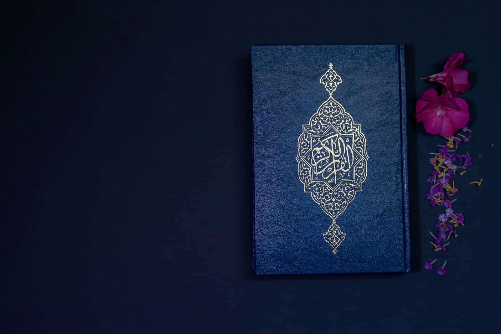 A Quran with gold calligraphy on a deep blue background, beside scattered flowers